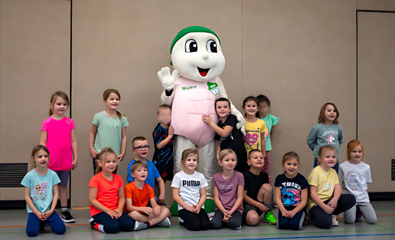 Eine Gruppe von 17 Grundschulkindern posiert in einer Turnhalle für ein Gruppenfoto. In ihrer Mitte steht ein großes, weißes Maskottchen mit grünem Kopf und rosa Oberteil, das freundlich in die Kamera winkt. Einige Kinder knien in der vorderen Reihe, während andere hinter ihnen stehen und lächeln.