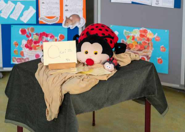 Ein großer, roter Marienkäfer-Stofftier-Charakter mit schwarzen Punkten und gelben Augen liegt auf einem dunkelgrün und beige drapierten Tisch. Vor dem Stofftier steht ein kleines, helles Schild mit handschriftlichem Text, der teilweise verdeckt ist. Rechts neben dem großen Stofftier liegt ein kleineres Marienkäfer-Stofftier. Im Hintergrund sind Pinnwände mit Kinderzeichnungen oder Plakaten zu sehen, die Körbe mit roten und gelben Früchten/Gemüse darstellen.